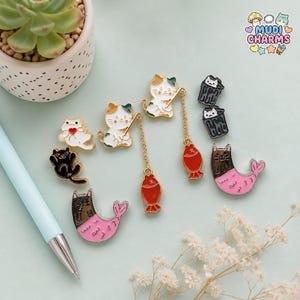 May include: A collection of enamel pins featuring cartoon cats, including two cat-mermaid hybrids with pink tails, two cats in trash cans, a cat holding a red heart, a black cat, and two cats fishing with dangling red fish charms on gold chains.