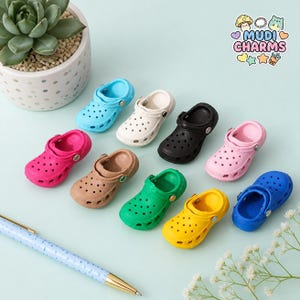 May include: A collection of nine miniature clog-style charms in various solid colors including blue, white, black, pink, tan, green, and yellow. Each charm features a perforated design and a pivoting heel strap. The text MUDI CHARMS is visible.