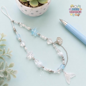 May include: A beaded phone charm featuring a white cord, clear and light blue beads, small silver-toned star charms, shell-shaped beads, and a translucent mermaid tail pendant. The charm includes a silver chain accent and a small pearl detail.