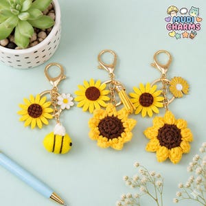 May include: Three sunflower keychains with gold-colored hardware. One keychain has a yellow and black bee charm, another has a yellow tassel, and the third has a small yellow flower charm. The keychains are on a light blue background.