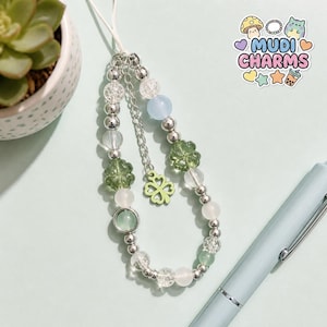 Pode incluir: Um pendente de telemóvel feito de missangas que combina contas metálicas prateadas, contas de vidro craquelado transparente e contas lisas em tons pastel verde, azul e branco. Inclui duas contas em forma de trevo e um pendente de trevo verde.