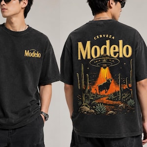 Może przedstawiać: Czarny t-shirt ze złotym napisem "Modelo" z przodu i pustynną sceną z bykiem i UFO z tyłu. Koszulka ma styl vintage.