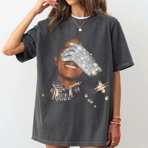 May include: Dark gray t-shirt featuring a portrait of a smiling person with a sparkling glove covering one eye. The image has a vintage look, with a black background and gold accents. The shirt is a casual, oversized style.