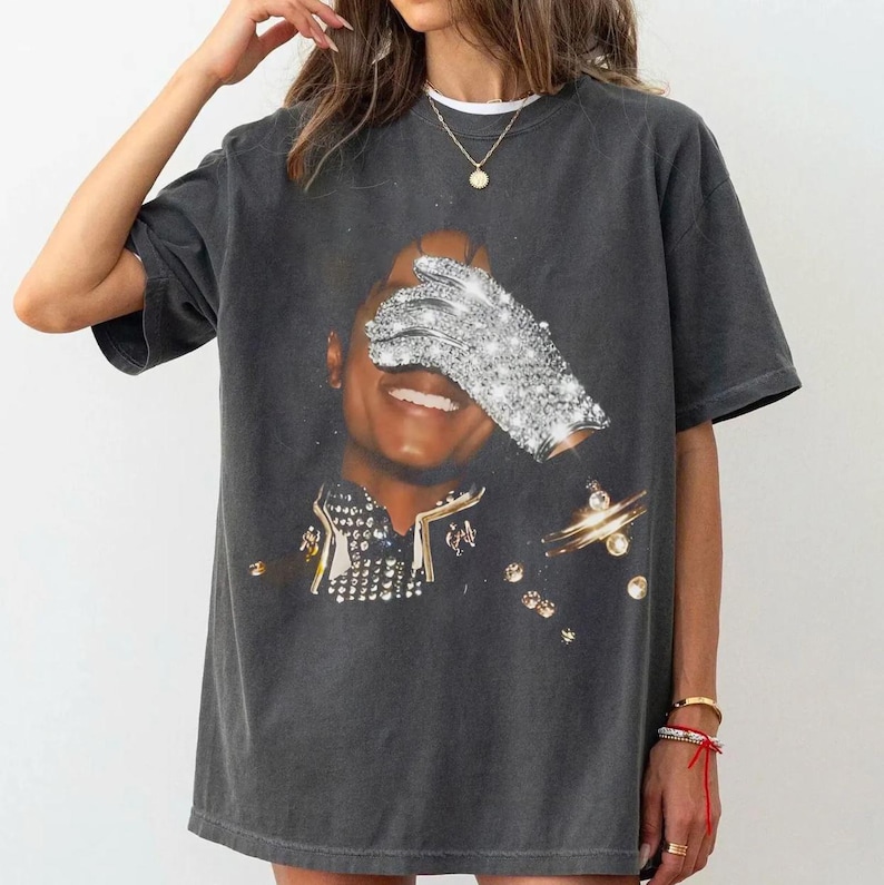 May include: Dark gray t-shirt featuring a portrait of a smiling person with a sparkling glove covering one eye. The image has a vintage look, with a black background and gold accents. The shirt is a casual, oversized style.