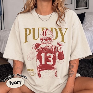 Könnte beinhalten: Ein elfenbeinfarbenes Kurzarm-T-Shirt mit einer verwaschenen rot-goldenen Grafik eines Footballspielers, der einen Helm und ein Trikot mit der Nummer 13 trägt. Der Schriftzug PUDY ist in großen goldenen Buchstaben über dem Spieler abgebildet.