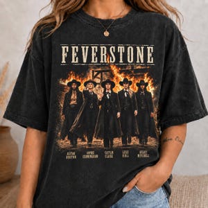 May include: A charcoal black mineral-washed t-shirt featuring a graphic print of five individuals in western attire standing before a large fire. The text FEVERSTONE is printed above the figures, with names listed below each person in the image.