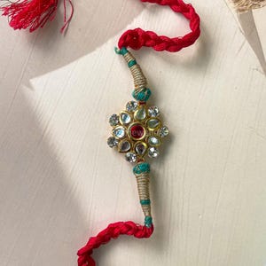 May include: A decorative wrist band featuring a central gold-tone floral motif set with clear teardrop-shaped stones and a single red circular stone. The band is made of braided red cord with gold and teal thread accents and a red tassel at one end.