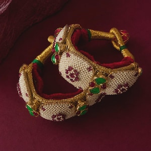 May include: A pair of ornate bangles featuring a gold-toned metal base wrapped in red fabric. The surface is decorated with white seed pearls, clusters of small red stones, and smooth green accents, creating a textured and layered appearance.