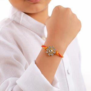 May include: A bracelet featuring a central gold-toned flower-shaped charm adorned with clear, sparkling rhinestones. The band is constructed from twisted orange and yellow threads, providing a vibrant contrast to the metallic centerpiece.