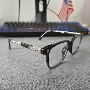 May include: Black eyeglasses with a rectangular frame and silver accents on the temples. The lenses are clear, and the frame has a small diamond-shaped detail near the nose bridge. The glasses are set against a gray surface.