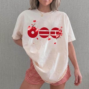 May include: Cream-colored t-shirt with a red graphic design featuring three connected circles and cherry blossom branches. The shirt is paired with red and white checkered shorts, creating a casual, spring-themed outfit.