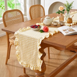 May include: A wooden dining table set with a yellow and white gingham table runner with ruffled edges. The table is set with a teapot, teacups, a bowl of berries, a peach, and a cookbook. Two wicker chairs are in the background.