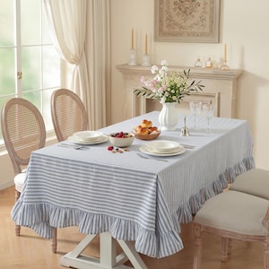 Könnte beinhalten: Ein Esstisch, gedeckt mit einer blau-weiß gestreiften Tischdecke mit Rüschenkante. Der Tisch ist mit weißen Tellern, Besteck und einer Schüssel mit Beeren gedeckt. Zwei Stühle mit Korbrücken sind zu sehen.