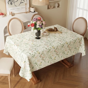 May include: A rectangular tablecloth with a floral pattern in shades of green, pink, and yellow covers a wooden table. The table is set with a vase of flowers, a cup of tea, and bread. Two chairs are visible.