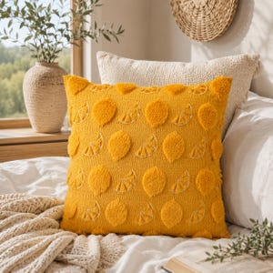 Könnte beinhalten: Ein quadratisches, gelbes Zierkissen mit einem Zitronenmuster. Das Kissen hat erhabene, strukturierte Zitronenformen und gestickte Details. Das Kissen liegt auf einem Bett mit anderen Kissen und einer gewebten Decke.