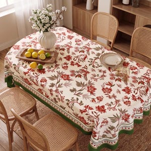 May include: An oval tablecloth with a floral pattern in red, green, and gray on an off-white background. The tablecloth is on a wooden table with wicker chairs. A vase of white flowers and a wooden tray with lemons and limes are on the table.