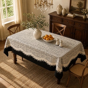 May include: A rectangular tablecloth with a black and white pattern covers a wooden table. The tablecloth has a black border with a white wavy design. A white vase with greenery and a bowl of oranges are on the table.