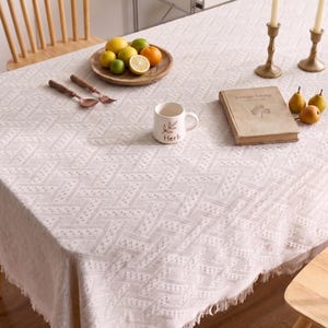 Könnte beinhalten: Ein weißes, strukturiertes Tischtuch mit geometrischem Muster bedeckt einen Tisch. Ein Holzbrett enthält Zitronen, Limetten und eine Orange. Eine Tasse mit der Aufschrift "Herb" befindet sich in der Nähe eines Buches und Birnen.