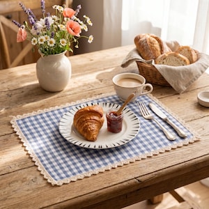 Könnte beinhalten: Frühstücksszene auf einem Holztisch. Ein Croissant und Marmelade auf einem Teller auf einer blau-weiß karierten Tischdecke. Eine Tasse Kaffee, Brot in einem Korb und eine Blumenvase vervollständigen das Arrangement.