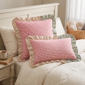 May include: Two decorative pillows with a pink and white gingham pattern. The pillows have ruffled edges in shades of beige, green, and pink. They are placed on a bed with a white comforter. A small side table with a mug and book is visible.
