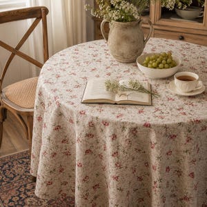 Könnte beinhalten: Eine runde Tischdecke mit einem sich wiederholenden Muster aus kleinen rosa Rosen und grünen Blättern auf cremefarbenem Grund. Der Stoff liegt auf einem runden Tisch, auf dem ein offenes Buch, eine weiße Schale mit grünen Trauben und ein Krug stehen.