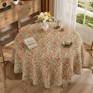 May include: A round tablecloth with a floral pattern in red, blue, and beige covers a wooden table. A vase of flowers, a book, and a cup of coffee sit on the table. The table is surrounded by wooden chairs.