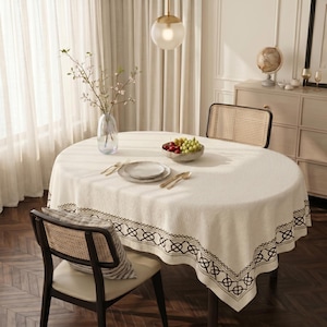May include: A round, cream-colored tablecloth with a geometric border design. The table is set with plates, silverware, and a bowl of fruit. A vase of flowers and a decorative globe are also visible.