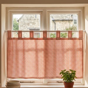 Könnte beinhalten: Ein rot-weiß karierter Küchenvorhang hängt vor einem Fenster. Der Vorhang wird von einer weißen Stange gehalten und hat ein schlichtes, klassisches Design. Eine kleine Topfpflanze steht auf der Fensterbank unter dem Vorhang.