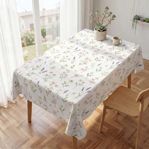 Könnte beinhalten: Ein rechteckiges, weißes Tischtuch mit einem floralen Muster aus rosa, gelben und lila Blumen und grünen Blättern. Das Tischtuch liegt auf einem Holztisch mit einem Holzstuhl. Eine Tasse und Topfpflanzen befinden sich ebenfalls auf dem Tisch.