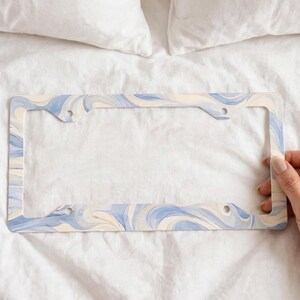 May include: A rectangular license plate frame featuring a swirling marbled pattern in shades of light blue, cream, and white. The frame has four circular mounting holes, one in each corner, and a smooth finish with a continuous fluid design.
