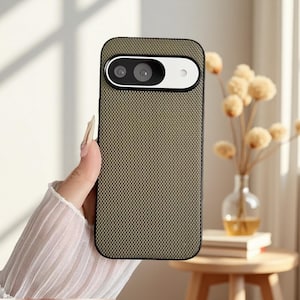 Può includere: Custodia per telefono verde oliva con motivo strutturato e bordo nero. La custodia presenta un ritaglio per la fotocamera con un accento bianco. La custodia del telefono è tenuta in mano.