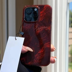 May include: A burgundy phone case with a textured pattern and the word "Liveshe" in gold script. The case has a black camera module and a blank white tag attached.