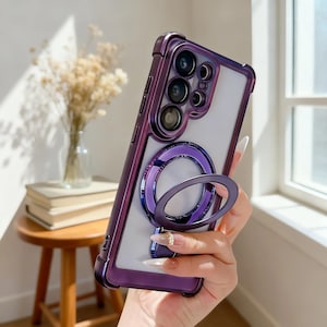 Könnte beinhalten: Violette Handyhülle mit durchsichtigem Rücken und integriertem Ringhalter. Die Hülle hat ein schützendes Stoßfängerdesign und eine metallisch-violette Oberfläche. Der Ringhalter ist ebenfalls violett und ermöglicht einen sicheren Halt oder Stand.