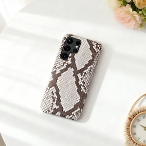 May include: A smartphone case with a snakeskin pattern in shades of brown and white. The case has a rectangular shape with a camera module on the top left corner. The case is lying on a white surface.