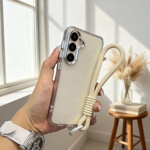 May include: Clear phone case with a cream-colored lanyard. The case has a transparent design with a silver camera module. The lanyard is made of a soft, woven material. The phone case is held in a hand.