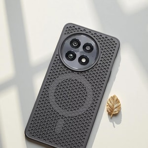 Puede incluir: Una funda de teléfono gris oscuro con un módulo de cámara circular y un diseño perforado. La funda tiene un acabado mate y un círculo magnético en la parte posterior. Una pequeña hoja dorada está al lado de la funda.