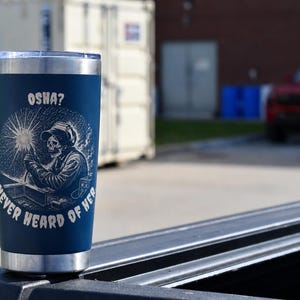 Puede incluir: Vaso aislado azul marino con borde plateado. El vaso presenta un gráfico blanco de un esqueleto soldador con el texto "OSHA? NEVER HEARD OF HER" en un diseño circular. El vaso está sobre la plataforma de una camioneta.