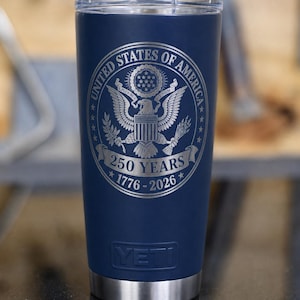 May include: A navy blue insulated tumbler with a silver rim and lid. The tumbler features the engraved United States of America seal with the text "250 YEARS 1776-2026". The brand name "YETI" is visible.