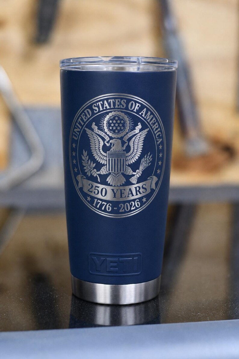 May include: A navy blue insulated tumbler with a silver rim and lid. The tumbler features the engraved United States of America seal with the text "250 YEARS 1776-2026". The brand name "YETI" is visible.
