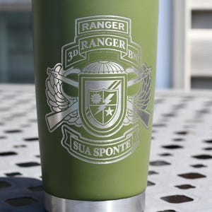 May include: Olive green insulated tumbler with a silver lid and base. The tumbler features a detailed silver emblem with the words "RANGER", "3D BN", and "SUA SPONTE". The design includes wings, guns, and a shield.