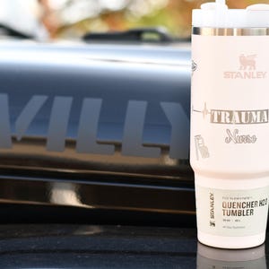 May include: A light pink Stanley Quencher H2.0 Tumbler with a white handle and lid. The tumbler has the word "STANLEY" and the text "TRAUMA Nurse" printed on it. The tumbler is sitting on a black surface.