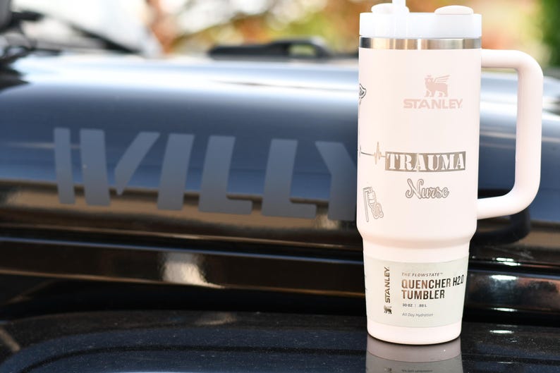 May include: A light pink Stanley Quencher H2.0 Tumbler with a white handle and lid. The tumbler has the word "STANLEY" and the text "TRAUMA Nurse" printed on it. The tumbler is sitting on a black surface.