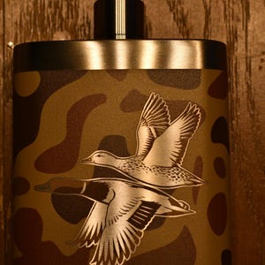 May include: A stainless steel flask featuring a camouflage pattern in shades of brown and tan. The front surface displays an engraved illustration of two ducks in flight, with the brand name YETI embossed in capital letters at the bottom.