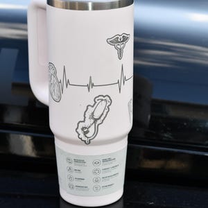 May include: A white insulated travel mug with a handle, decorated with medical symbols. The design includes a heartbeat line, a caduceus, a stethoscope, and an anatomical heart. The bottom of the mug has icons and text.