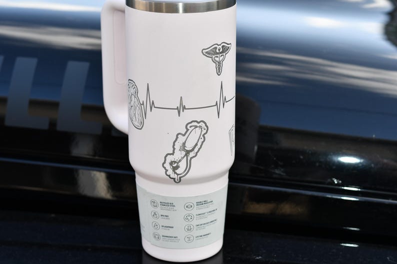 May include: A white insulated travel mug with a handle, decorated with medical symbols. The design includes a heartbeat line, a caduceus, a stethoscope, and an anatomical heart. The bottom of the mug has icons and text.