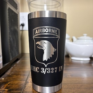 May include: A matte black stainless steel tumbler featuring a silver laser-engraved design of the 101st Airborne Division insignia. The graphic includes the word AIRBORNE at the top, an eagle head in the center, and the text HHC 3/327 IN below.