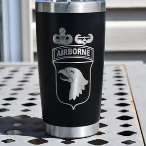 May include: Black insulated tumbler with a silver lid and base. The tumbler features a silver emblem with the word "AIRBORNE" and a shield with an eagle head. The tumbler is designed to keep beverages hot or cold.