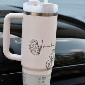 May include: A pale pink insulated travel mug with a white lid, handle, and straw. The mug has a heart and heartbeat line graphic. The mug is designed to keep drinks cold or hot.