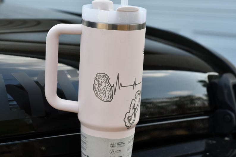 May include: A pale pink insulated travel mug with a white lid, handle, and straw. The mug has a heart and heartbeat line graphic. The mug is designed to keep drinks cold or hot.