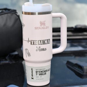 May include: A light pink Stanley Quencher H2.0 Flowstate tumbler with a white handle and lid. The tumbler has the word "TRAUMA Nurse" and a heartbeat graphic. The tumbler also has the Stanley logo and the words "Quencher H2.0 Tumbler" printed on it.
